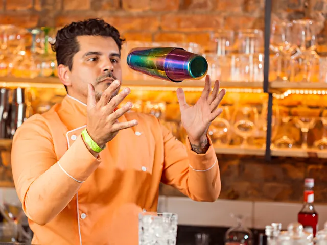 A professional bartender tosses a colorful cocktail shaker in a vibrant bar setting.