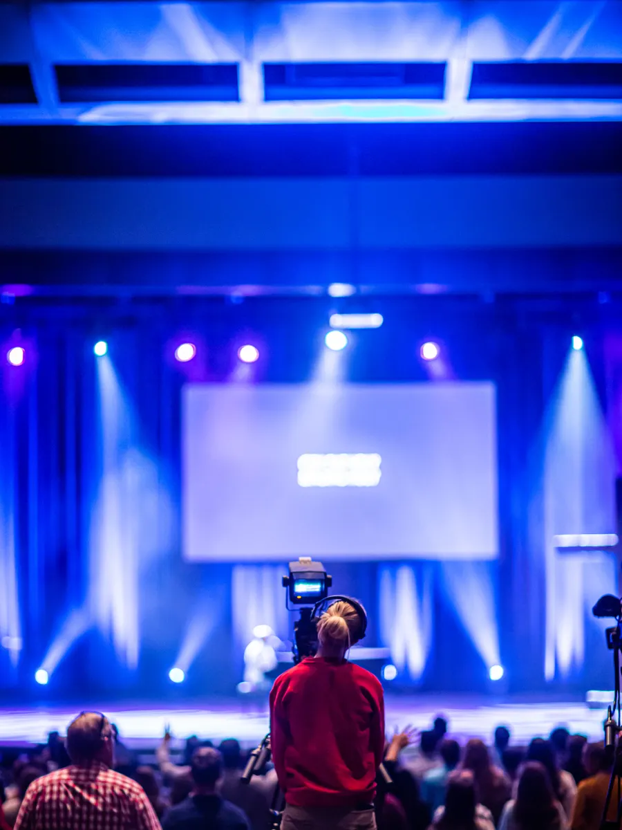 Cameraman capturing a live event on stage with colorful lights and blurred audience.