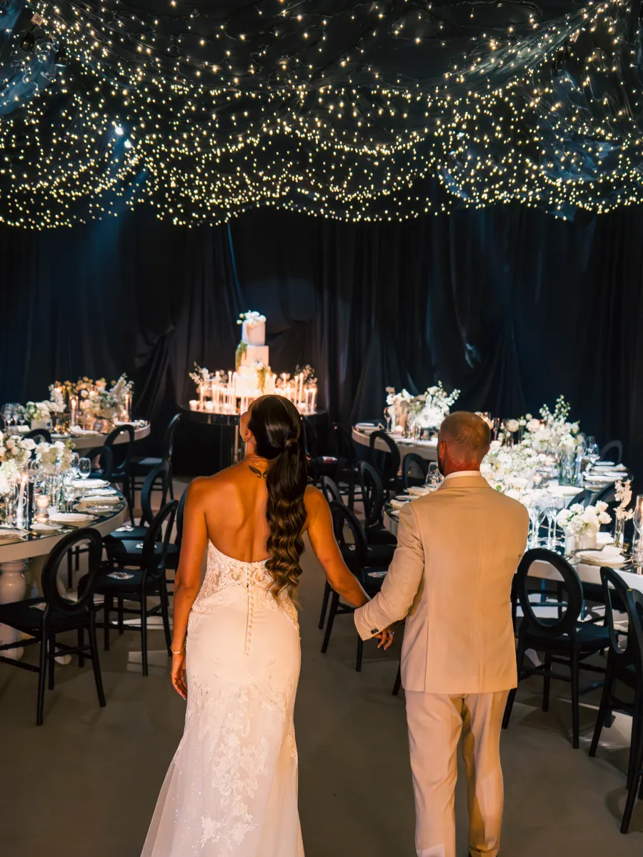 A couple entering a beautifully decorated indoor wedding reception with string lights and elegant decor.